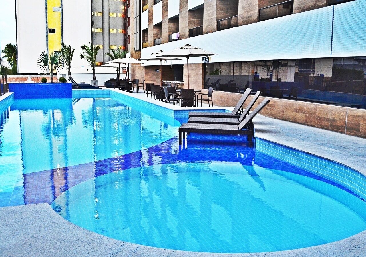 Una piscina con sillones de descanso y sombrillas, rodeada por un edificio con fachada de azulejos.