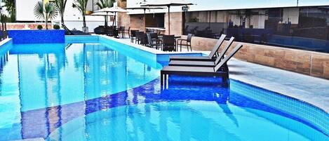 Una piscina con sillones de descanso y sombrillas, rodeada por un edificio con fachada de azulejos.