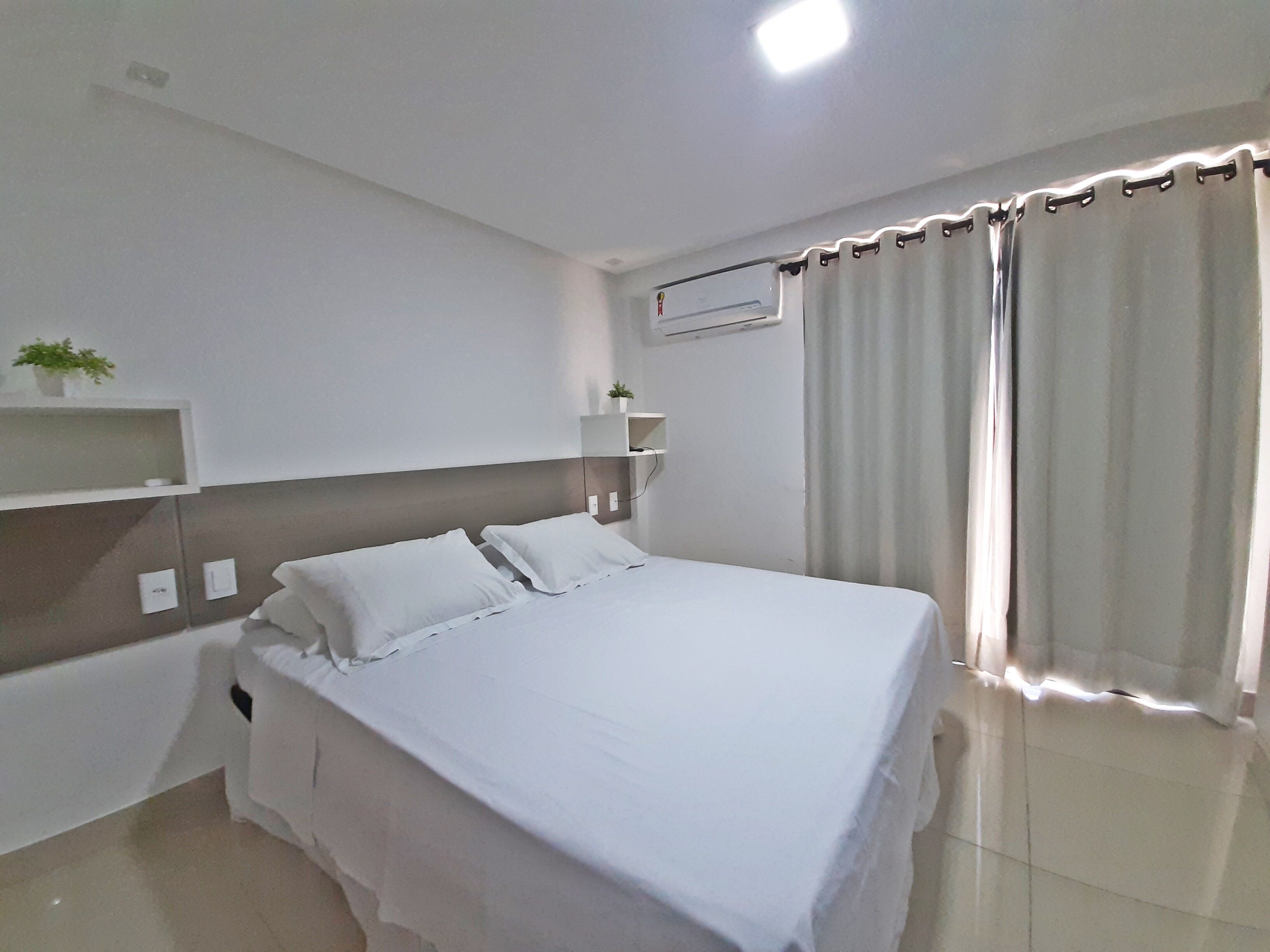 A modern hotel room with a large bed, a bedside table, a shelf with a plant, and a window with curtains.