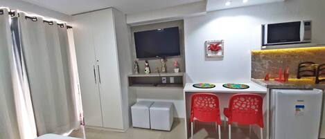 A compact hotel room with a bed, a desk with two chairs, a small refrigerator, and a TV mounted on the wall.