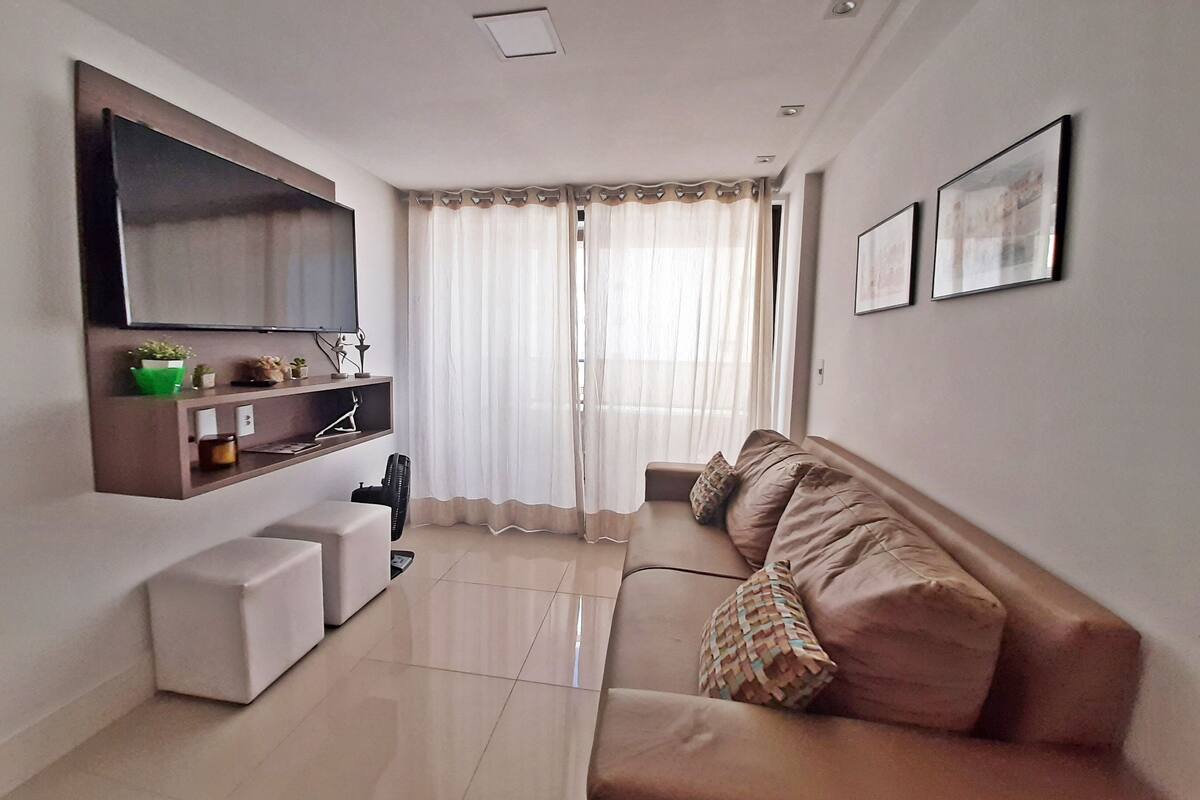 Una sala moderna con un televisor de pantalla plana montado en la pared, un sofá, una mesita de centro y una ventana con cortinas traslúcidas.