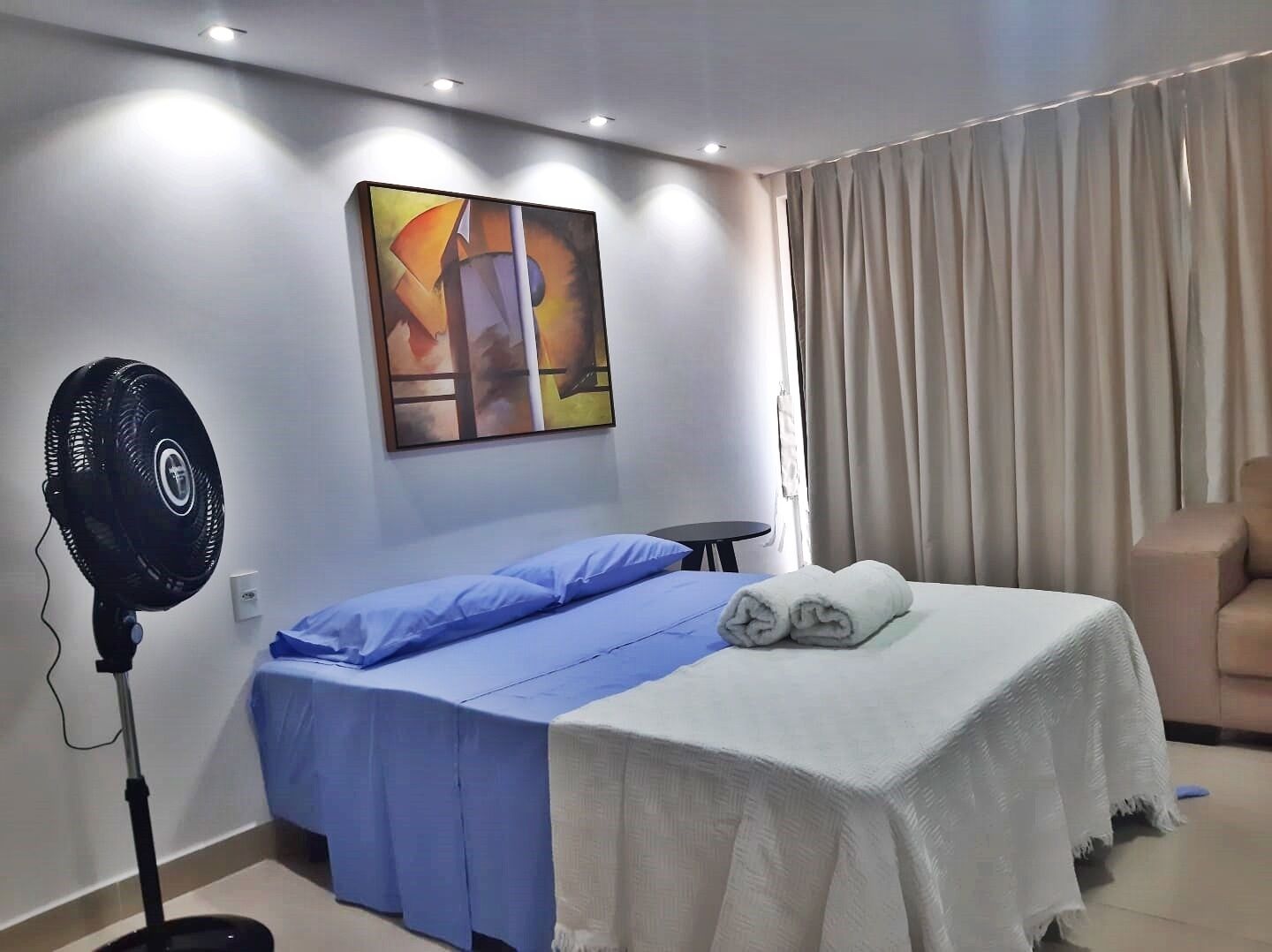 Una habitación de hotel con cama, ventilador, mesa, silla y un cuadro en la pared.