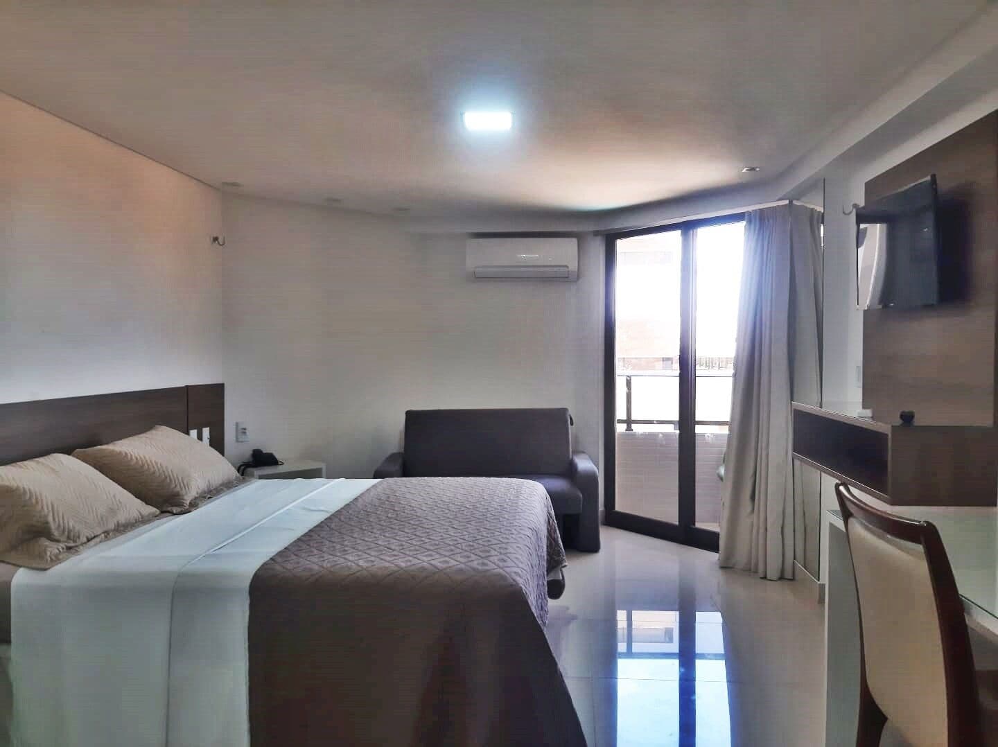 Quarto de hotel com cama, escrivaninha, cadeira, televisão e porta de vidro corrediça para uma varanda.