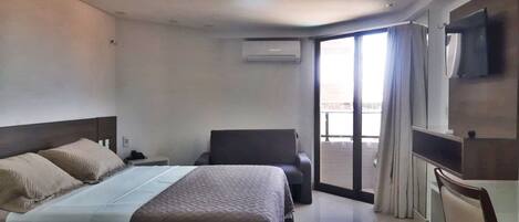 Quarto de hotel com cama, escrivaninha, cadeira, televisão e porta de vidro corrediça para uma varanda.