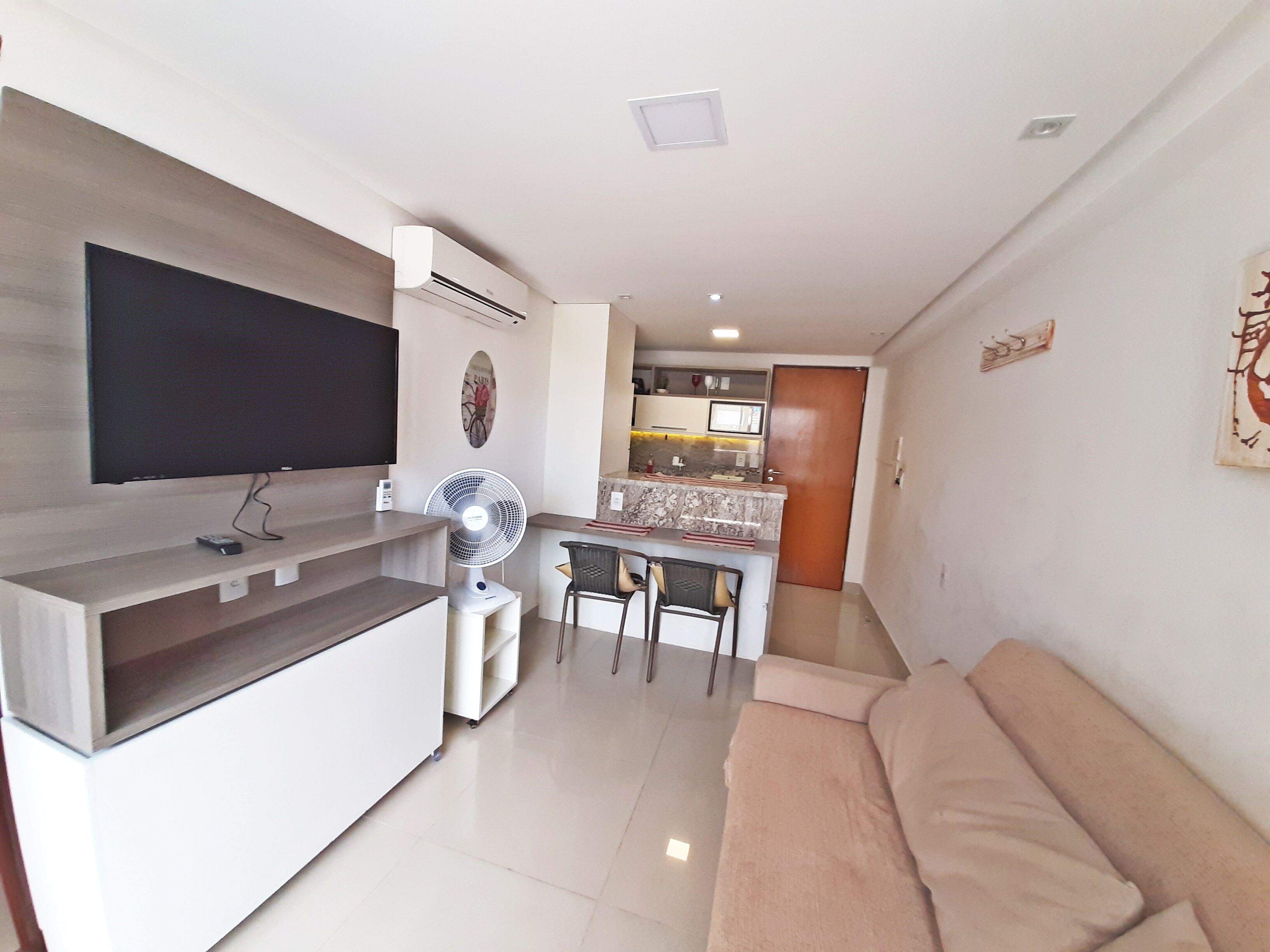 A modern living room with a flat-screen TV, a standing fan, a sofa, and a kitchen area in the background.