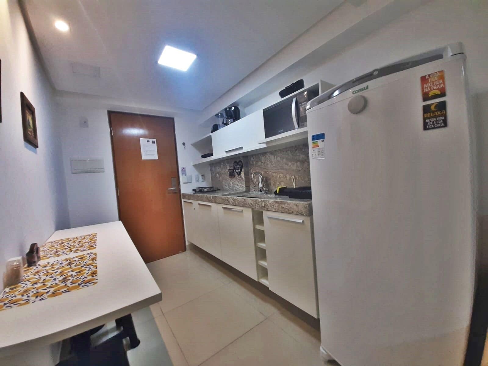 A compact kitchen with white cabinets, a countertop, and a refrigerator. There is a door with a sign, a small sink, and a microwave on the countertop.