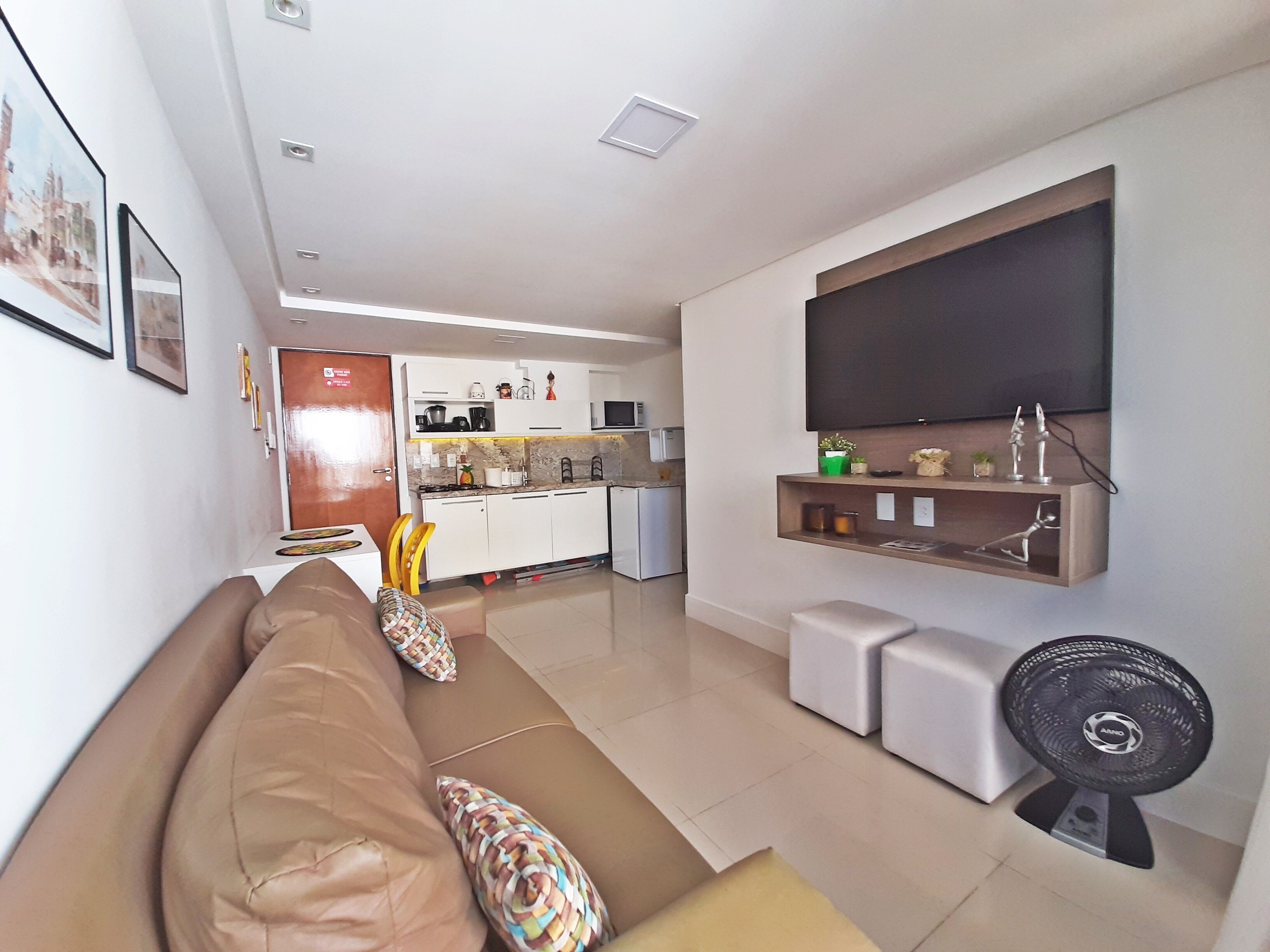 A modern living room with a sofa, a flat-screen TV mounted on the wall, and a kitchen in the background.