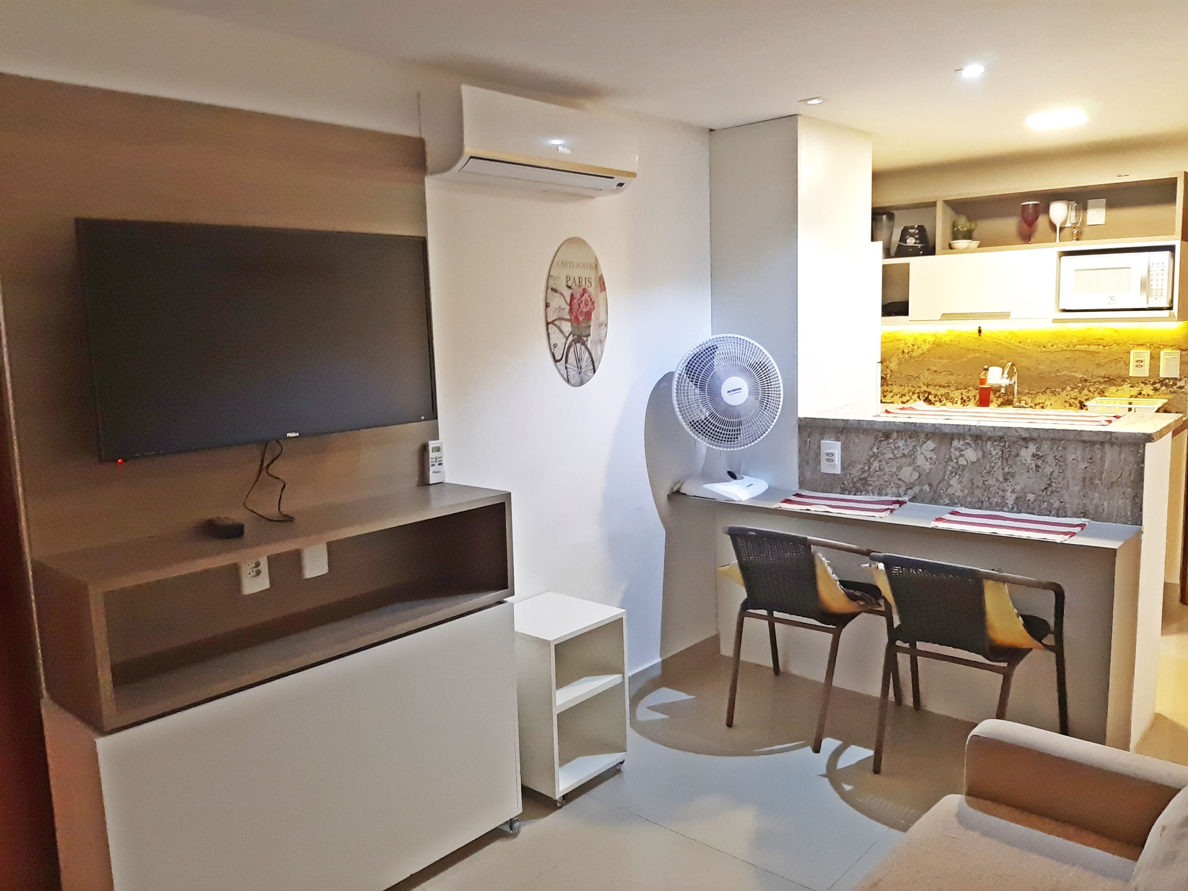 Un salon moderne doté d’un téléviseur à écran plat, d’un ventilateur sur pied, d’une petite armoire blanche et d’un coin repas avec deux chaises.