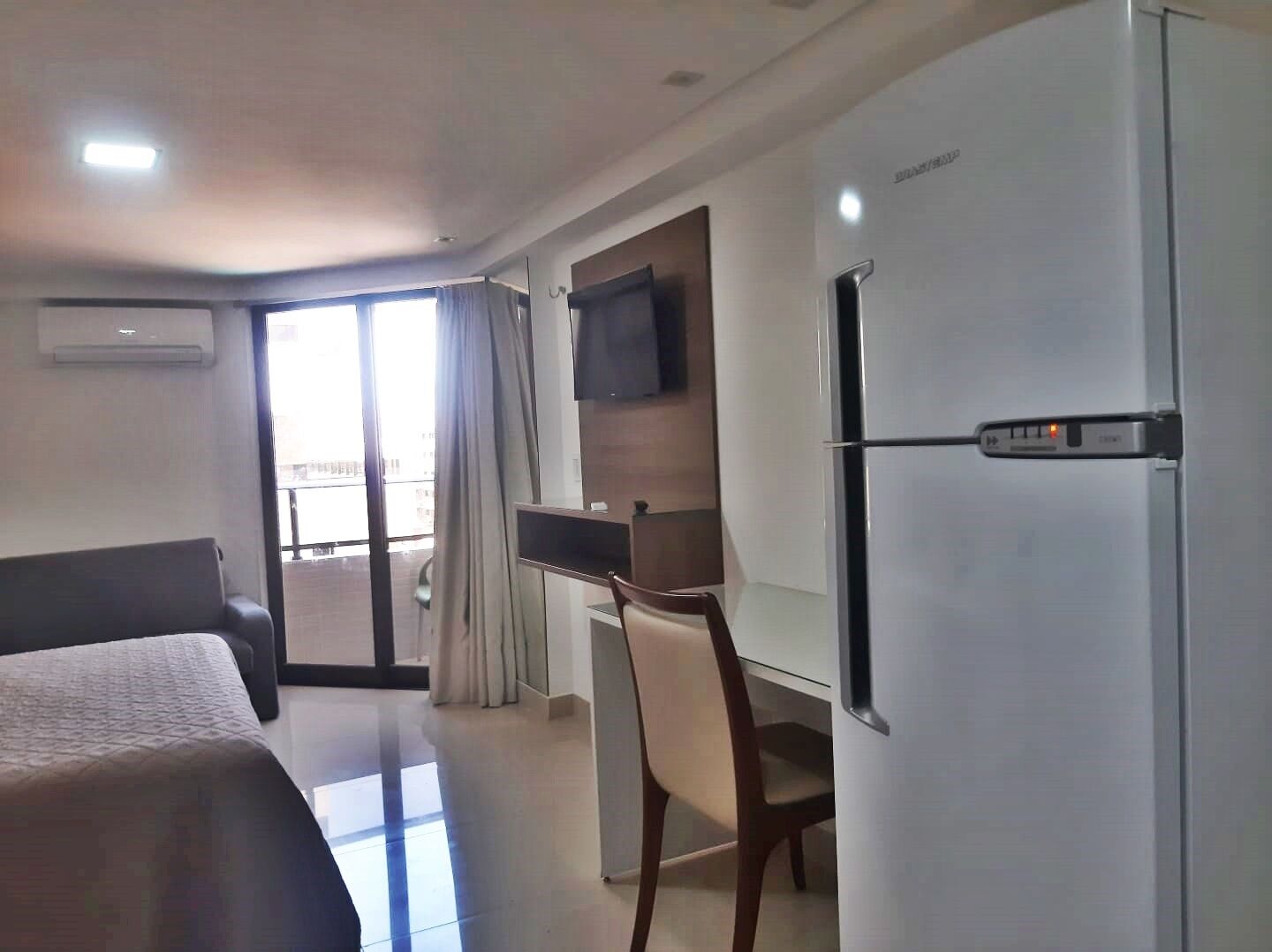 A hotel room with a bed, a desk, a chair, a TV mounted on the wall, and a refrigerator.