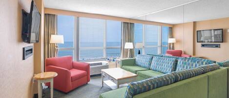 Habitación de hotel con un ventanal grande, un sofá verde, un sillón rojo, una mesita y un televisor montado en la pared.
