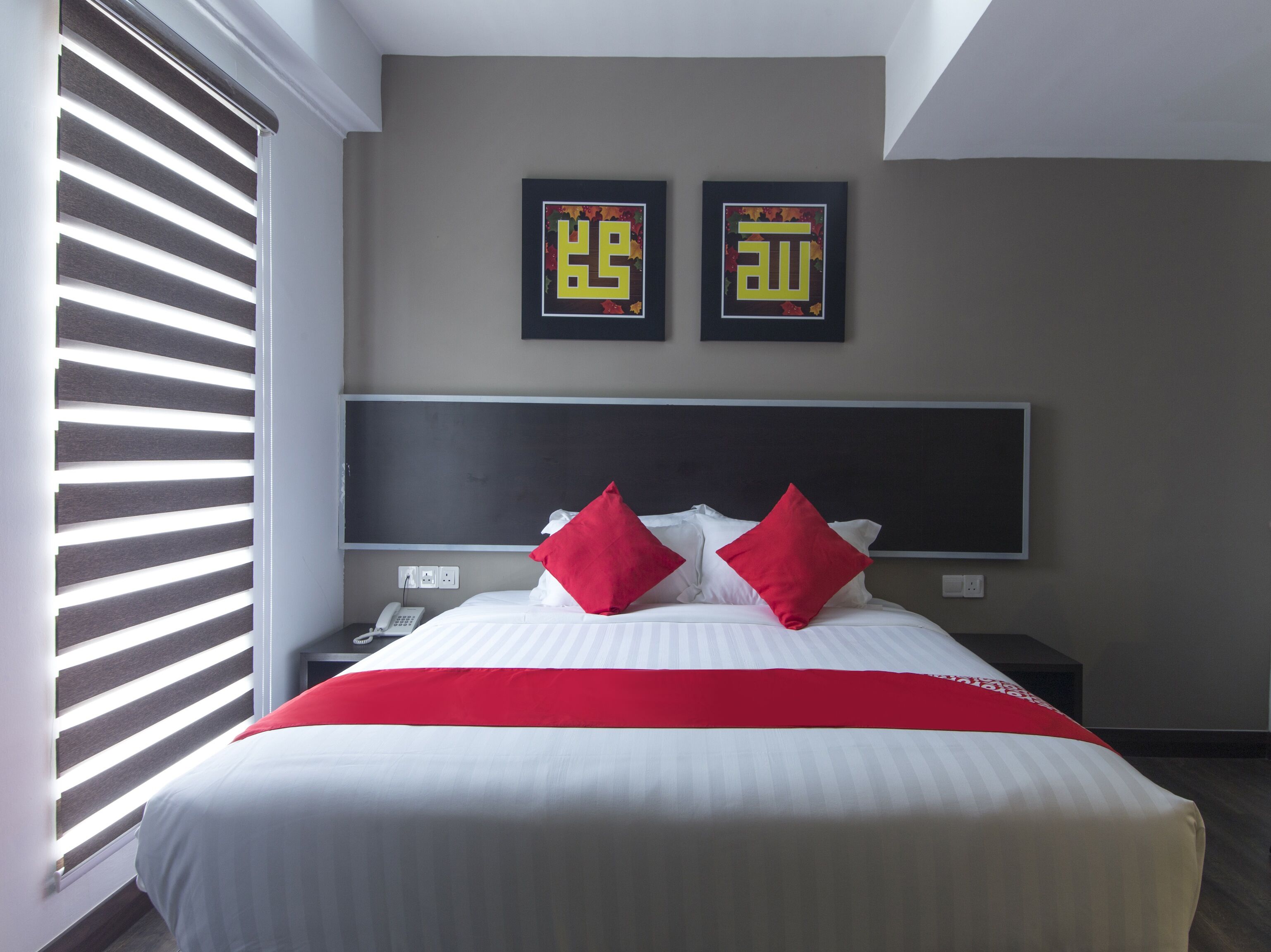 A hotel room with a large bed, red pillows, a bedside table with a phone, and two framed calligraphy pieces on the wall.