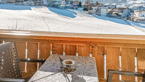 A wooden deck with a table and chairs, overlooking a snowy landscape with buildings and mountains in the distance.