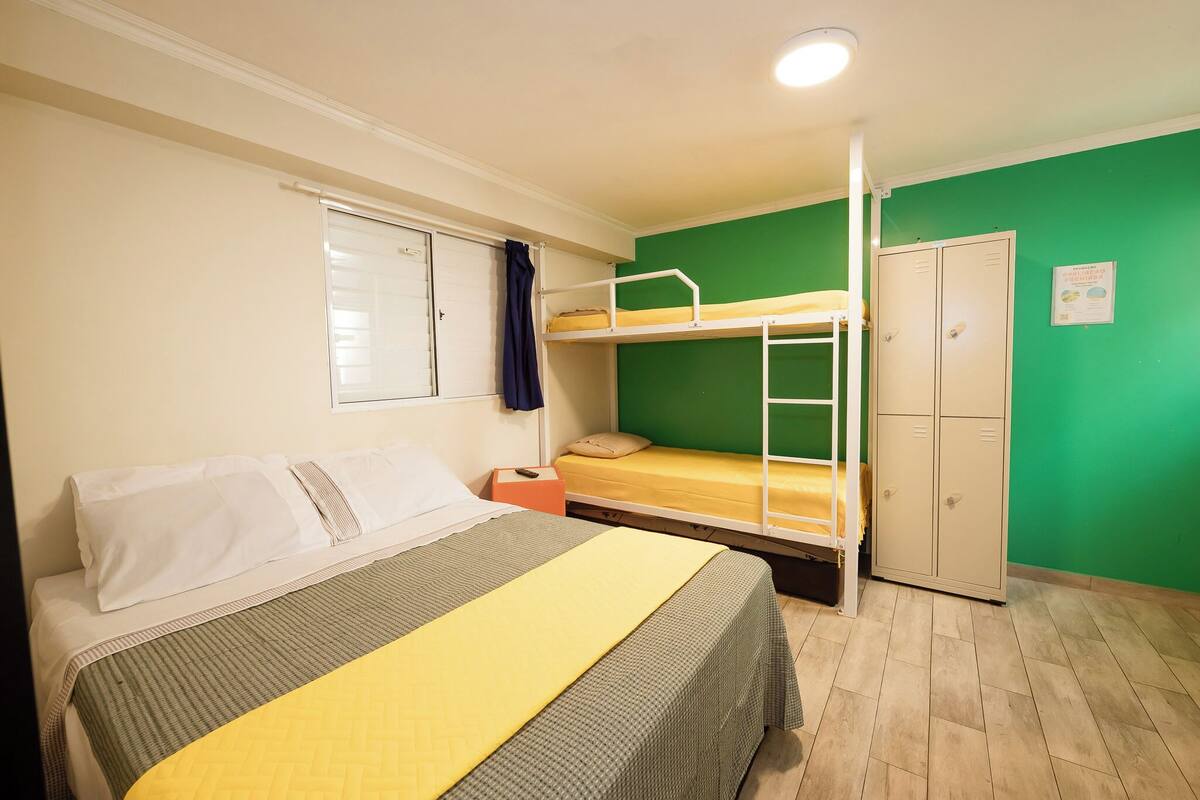 A dormitory room with bunk beds, a single bed, a wardrobe, and a window with curtains.