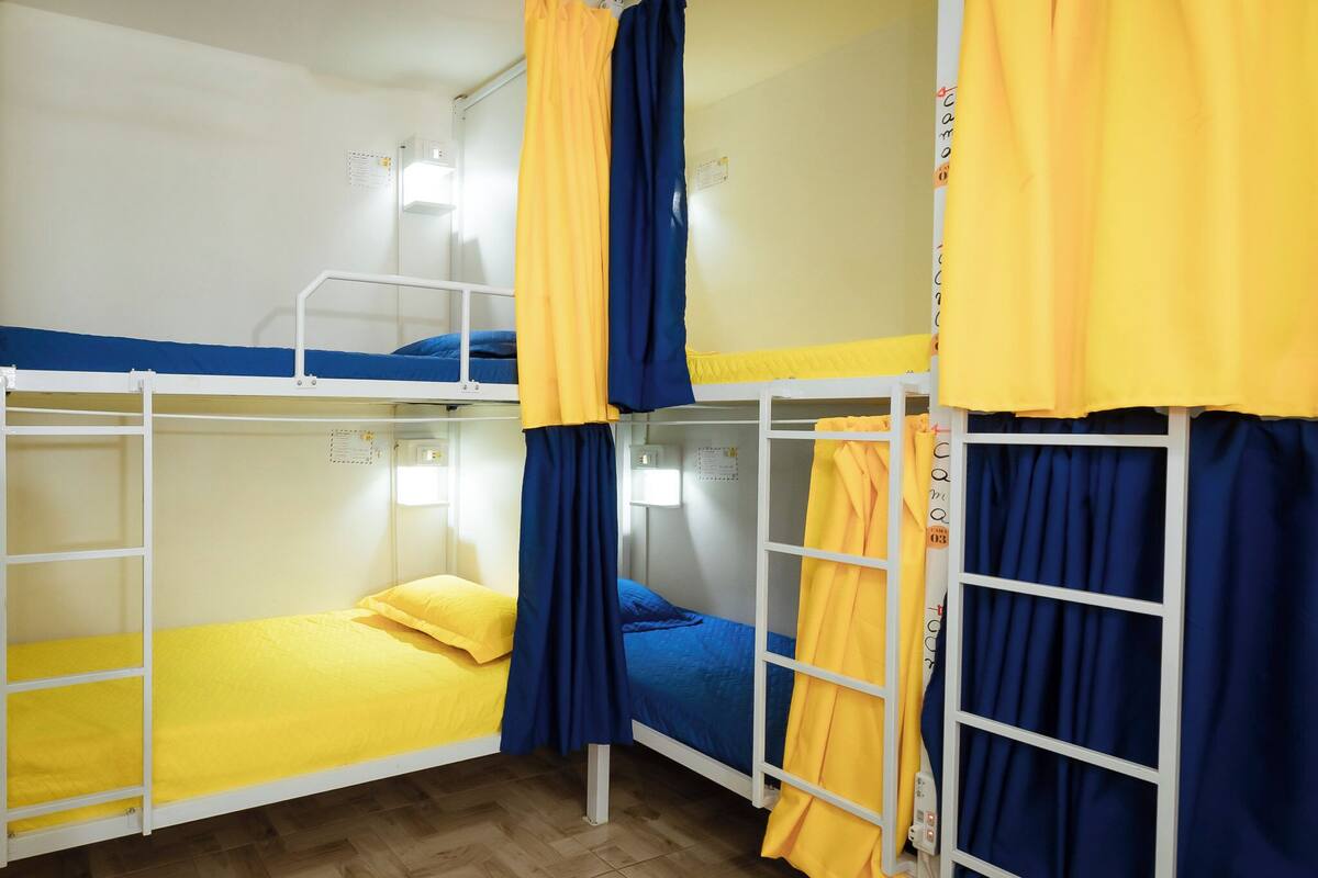 A dormitory room with bunk beds, each with a mattress and pillows, and curtains.