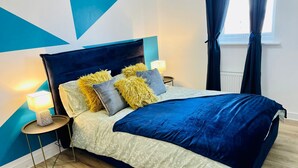 A modern bedroom with a geometric wall design, a large bed with a dark headboard, and a nightstand with a lamp.