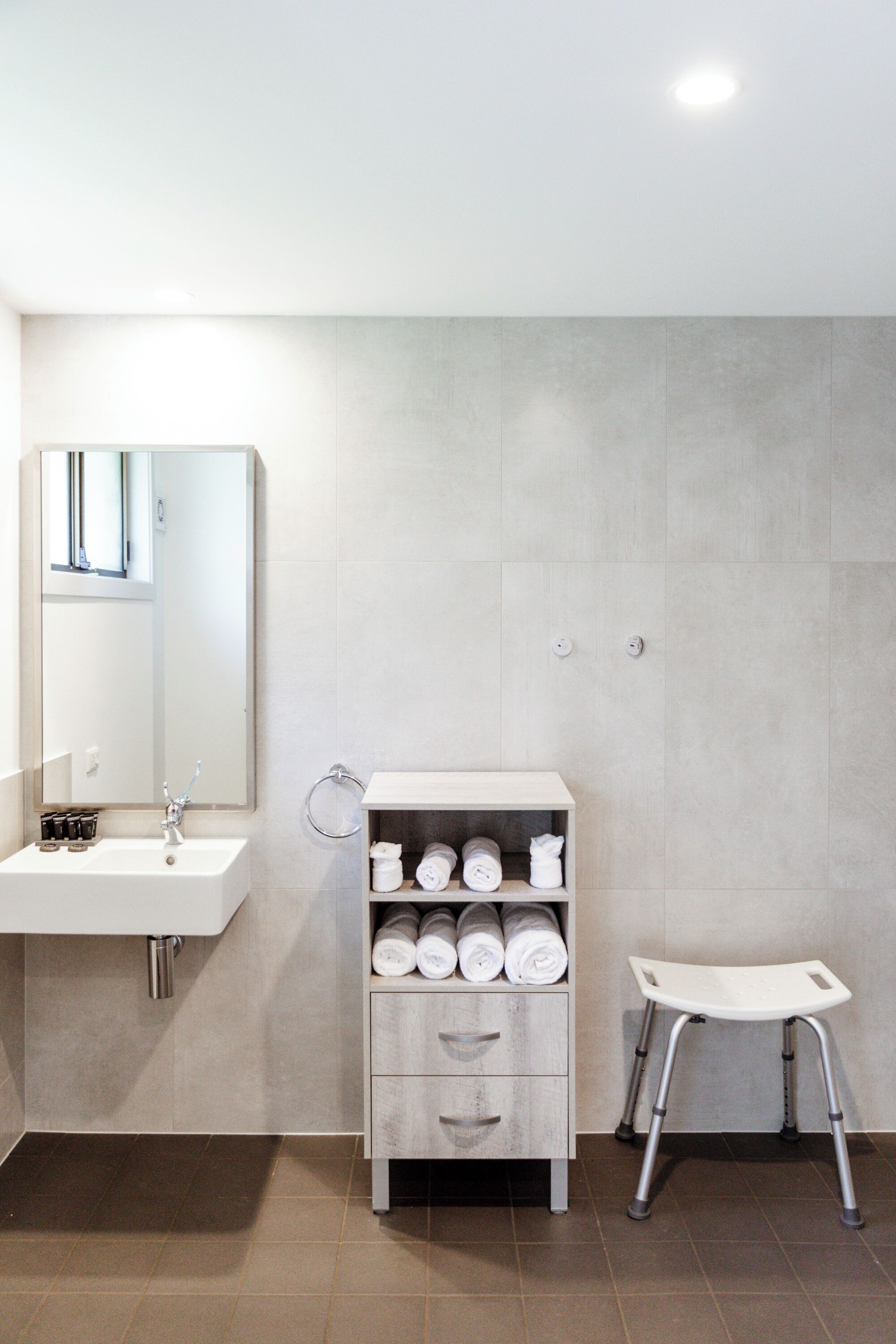 A modern bathroom with a white sink, a mirror, a towel rack, a storage cabinet, and a small stool.