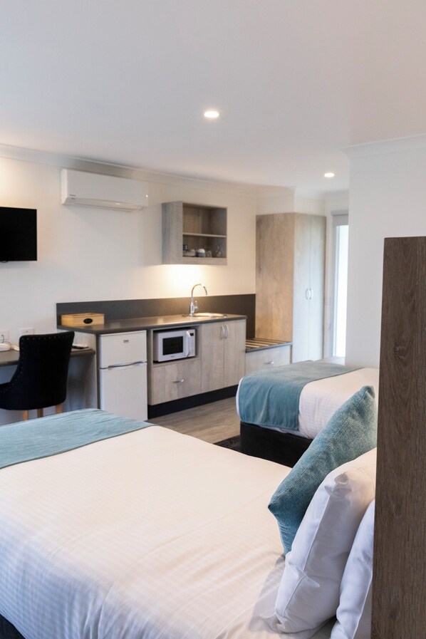 A modern hotel room with two beds, a kitchenette, a desk, and a TV.