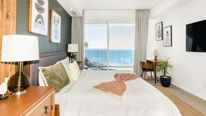 A hotel room with a large bed, a desk with a lamp, a chair, a TV, and a balcony with a view of the sea.