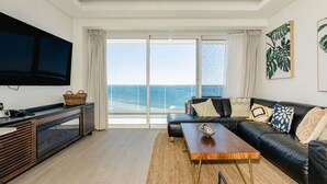A modern living room with a large flat-screen TV, a black leather sofa, a wooden coffee table, and a view of the ocean through floor-to-ceiling windows.