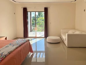 A hotel room with a bed, a white sofa, a round ottoman, and a sliding glass door leading to an outdoor area.