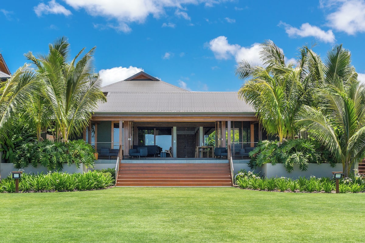 A modern house with a covered porch, surrounded by lush greenery and palm trees.