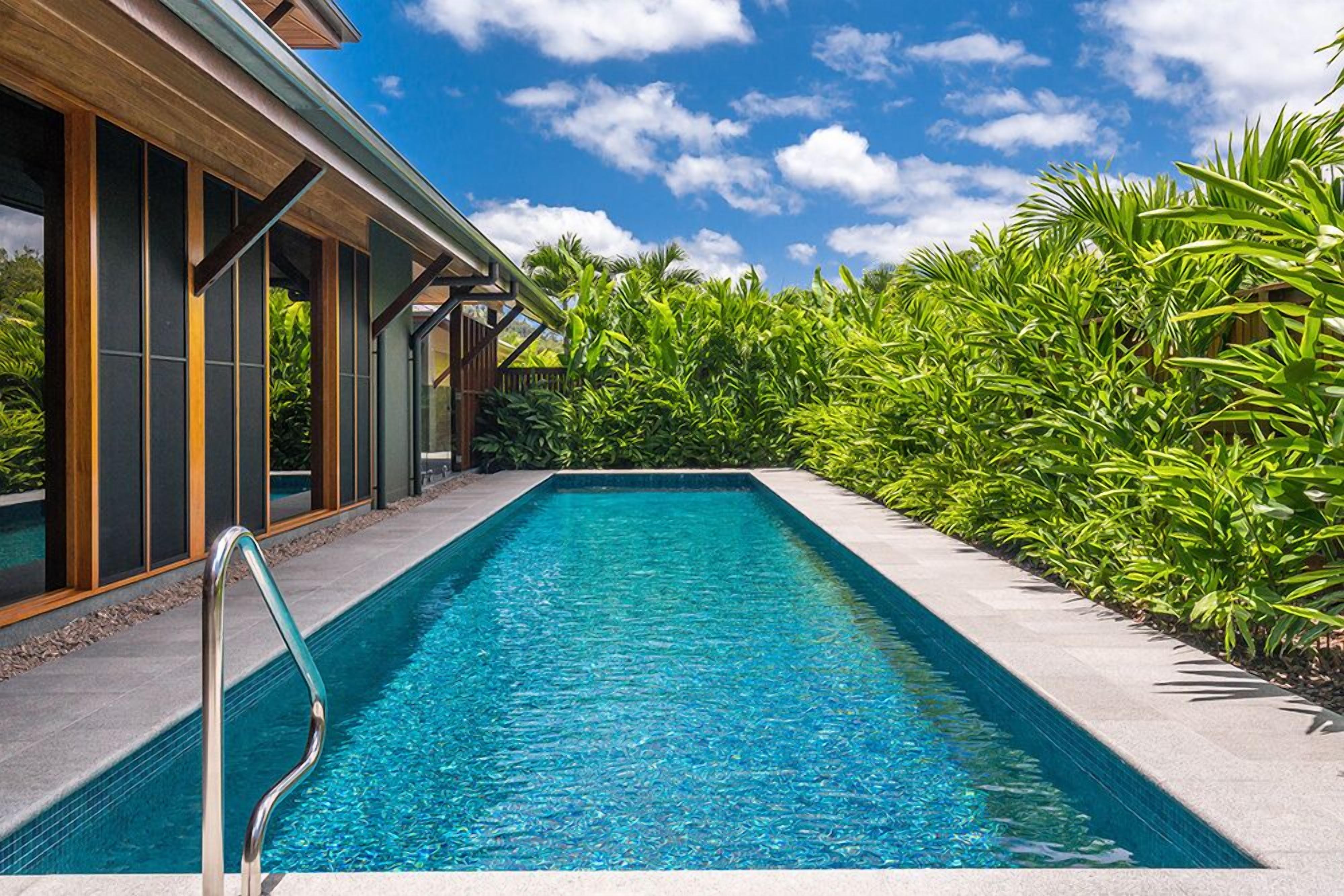 A rectangular swimming pool with a metal ladder, surrounded by lush greenery and a modern building with large windows.