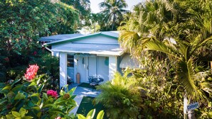 Una cabaña blanca con techo verde, rodeada de exuberante vegetación tropical y palmeras.