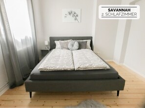 A bedroom with a grey bed, a nightstand, a lamp, and a framed picture on the wall.