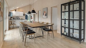 A modern kitchen with wooden cabinets, a dining area with a long table and chairs, and a black cabinet with glass doors. - 3 bedroom beautiful home in Aakirkeby (Aakirkeby)