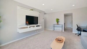 A modern living room with a flat-screen TV mounted on a white wall, a small table with a basket, and a potted plant. - Beautiful, Stylish 3 Bedroom Home in San Jose! (San Jose)