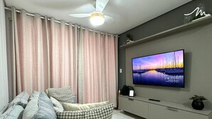 Una sala de estar moderna con un sofá gris, cojines estampados, un televisor de pantalla plana, un ventilador de techo y un estante decorativo.