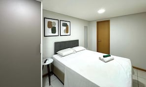 Quarto de hotel com uma cama, uma mesinha com um controle remoto, duas obras de arte abstratas emolduradas e uma porta de madeira.