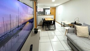 Una sala de estar moderna con un televisor de pantalla plana, un sofá gris, una mesa de comedor de madera y una cocina al fondo.