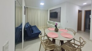 Een moderne woonkamer met een eethoek, een blauwe bank, een flatscreen televisie en een groot raam met gordijnen.