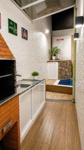 A kitchen with a brick oven, a sink, a potted plant, a stone wall, and a tiled pool area. - Beautiful house with a swimming pool heated by solar power year-round, near the beach. (Praia Grande)