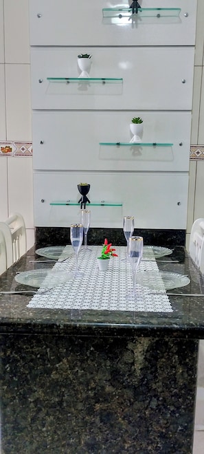 A dining table with a glass top, two wine glasses, a small potted plant, and a decorative item on a patterned table runner.