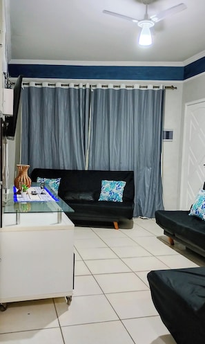 A living room with a black sofa, a glass coffee table, a ceiling fan, and a grey curtain.