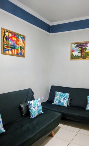A room with two black sofas, one with a blue and white patterned pillow, and a painting of a colorful park scene on the wall.
