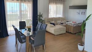 A living room with a sofa, a dining table with chairs, and a coffee table. There is a plant on the coffee table and another plant near the window. - Noza Beach (Valle-Niza)