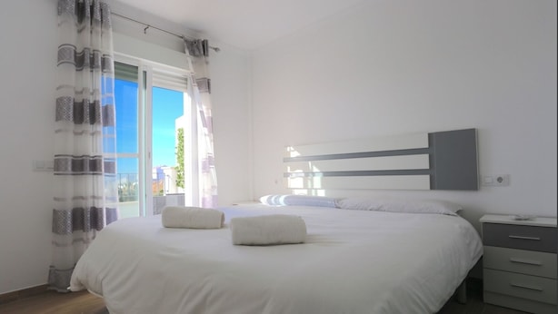 A modern bedroom with a large bed, a bedside table, a wardrobe, and a view of the outdoors. - Noza Beach (Valle-Niza)