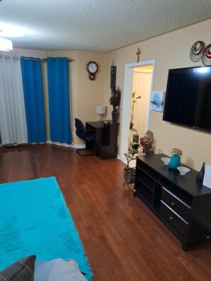 A living room with a wooden floor, a blue rug, a desk with a chair, a television, and a clock on the wall. - Luxury getaway (Brampton)