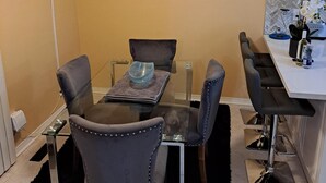 A dining area with a glass table, four chairs, a black rug, a painting of flowers, a TV mounted on the wall, and a kitchen area with a white countertop and a water bottle. - Luxury getaway (Brampton)