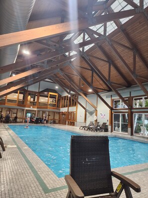 Une piscine intérieure dotée d’un plafond haut et de poutres en bois, entourée de chaises longues et recouverte d’un sol carrelé.