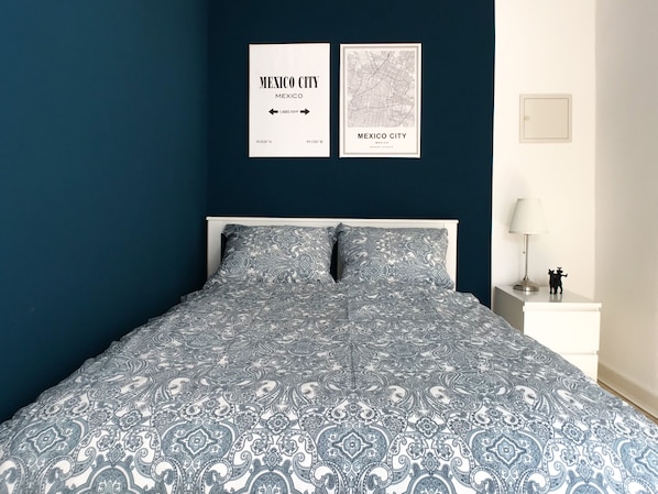 A bed with a patterned bedspread, a white nightstand with a lamp, and two framed maps on the wall.