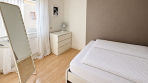 A bedroom with a bed, a nightstand, a mirror, a window with curtains, and a wall-mounted fan.