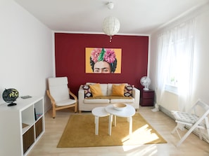 A living room with a red accent wall, a white sofa with patterned cushions, a white side table, a small white table with a bowl, a white chair, a white floor lamp, and a large window with sheer curtains.