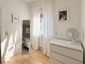 Une chambre à coucher avec un lit, une commode, un radiateur et une fenêtre ornée de rideaux légers.