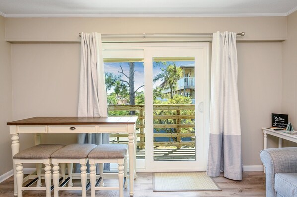 A room with a dining table, two chairs, a small desk, and a view through a glass door to an outdoor balcony.