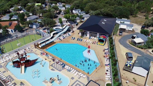 Una gran piscina con toboganes y una zona de juegos con temática de naufragio, rodeada de tumbonas y un área de estar cubierta.