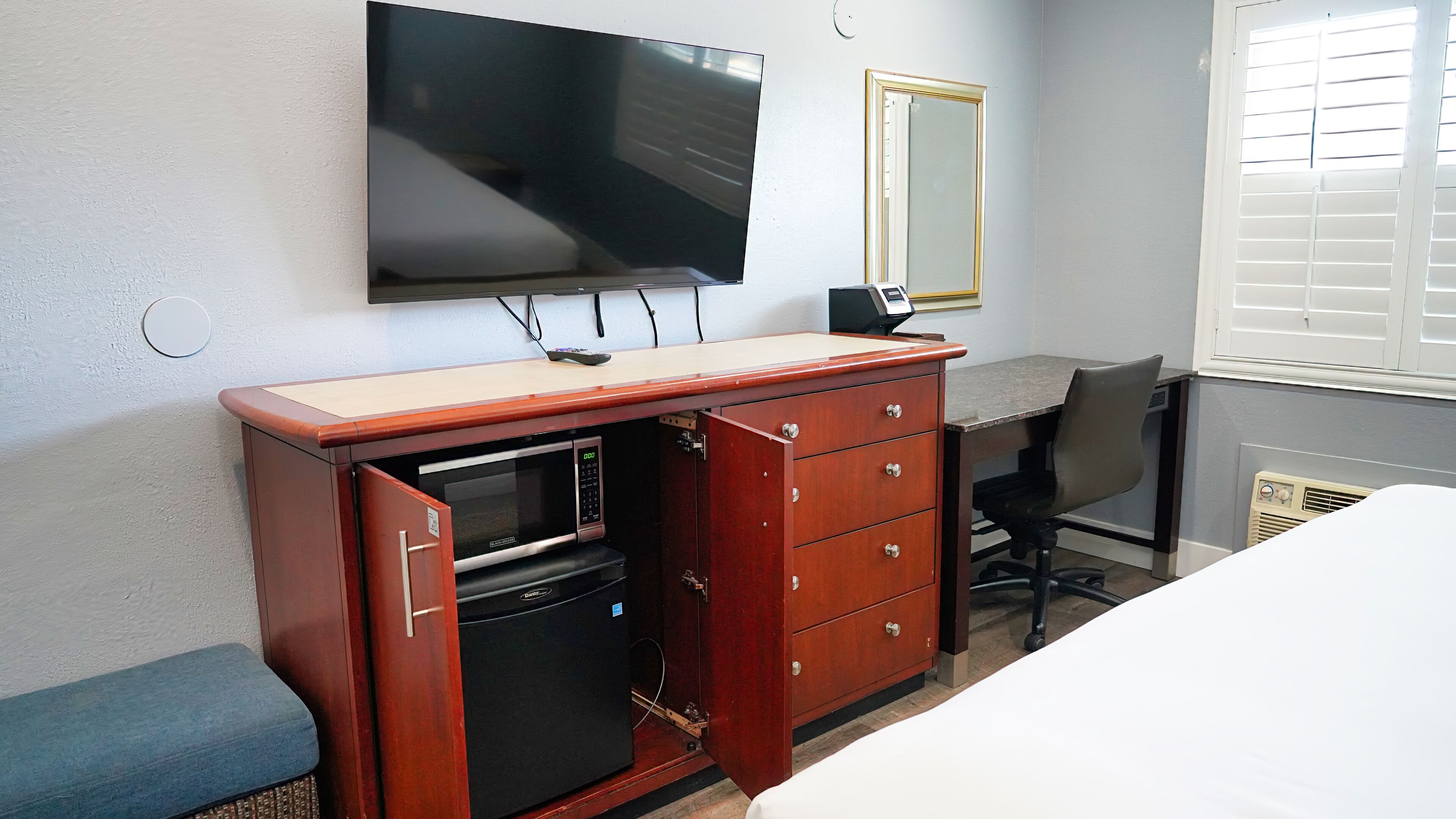 Une chambre d’hôtel équipée d’un téléviseur à écran plat fixé au mur, d’une commode en bois avec un micro-ondes, d’un bureau avec une chaise et d’une fenêtre avec des stores.